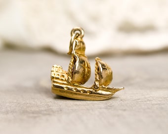May include: A small, gold-colored charm in the shape of a sailing ship. The charm features a detailed design of the ship's hull, sails, and rigging. The charm has a loop at the top for attaching to a necklace or bracelet. The charm is likely made of gold or gold-plated metal.