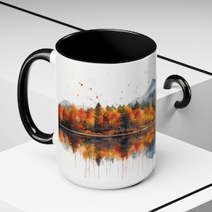 May include: White ceramic mug with a black handle and a black rim. The mug features a watercolor painting of a forest reflected in a lake. The trees are in shades of orange, yellow, and red, suggesting autumn.