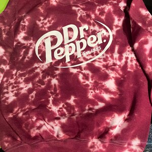 May include: A maroon tie-dye sweatshirt with the Dr Pepper logo in white. The logo includes the words "Dr Pepper" in a stylized font, with "Est. 1885" below. The sweatshirt has a crew neck and long sleeves.