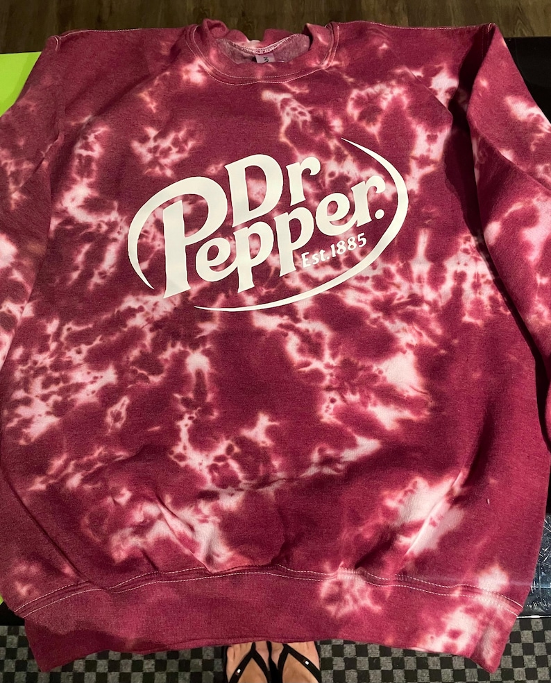 May include: A maroon tie-dye sweatshirt with the Dr Pepper logo in white. The logo includes the words "Dr Pepper" in a stylized font, with "Est. 1885" below. The sweatshirt has a crew neck and long sleeves.