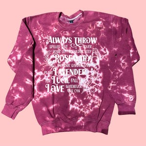 May include: A maroon tie-dye sweatshirt with white text. The text reads "ALWAYS THROW SPILLED SALT OVER YOUR LEFT SHOULDER KEEP ROSEMARY BY YOUR GARDEN GATE PLANT LAVENDER FOR LUCK FALL IN LOVE WHENEVER YOU CAN."