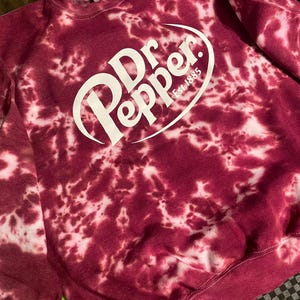 May include: A burgundy tie-dye sweatshirt with the Dr. Pepper logo in white. The logo includes the words "Dr Pepper" in a stylized font, with "Est 1885" below. The sweatshirt has a crew neck and long sleeves.