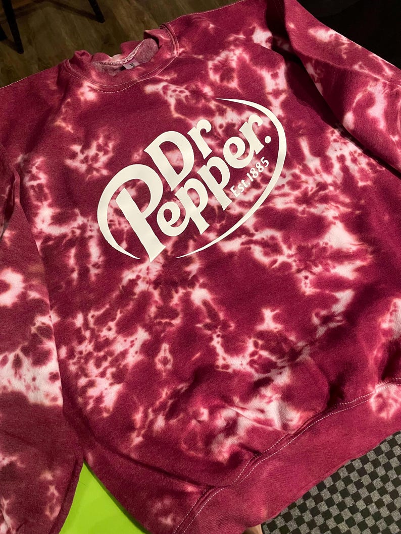 May include: A burgundy tie-dye sweatshirt with the Dr. Pepper logo in white. The logo includes the words "Dr Pepper" in a stylized font, with "Est 1885" below. The sweatshirt has a crew neck and long sleeves.