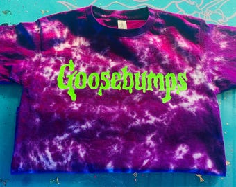 May include: A short-sleeved t-shirt featuring a purple and white tie-dye pattern. The word Goosebumps is printed across the chest in a bright lime green font with a distinct, jagged, and stylised lettering style.