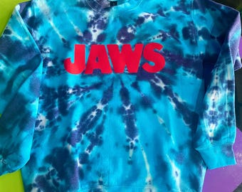 Op de afbeelding: Een blauwe tie-dye sweatshirt met het woord "JAWS" in grote rode letters over de borst. De sweatshirt heeft een ronde hals en lange mouwen. Het tie-dye patroon heeft tinten blauw en wit.