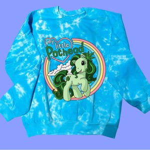 May include: A blue tie-dye sweatshirt featuring a cartoon pony with green hair and a cannabis leaf. The text "my little Pothead" is printed above the pony in a heart-shaped design, surrounded by a rainbow.
