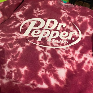 May include: Maroon tie-dye sweatshirt featuring the Dr Pepper logo in white. The logo includes the brand name and "Est. 1885". The sweatshirt has a crew neck and long sleeves. The fabric has a unique, mottled pattern.