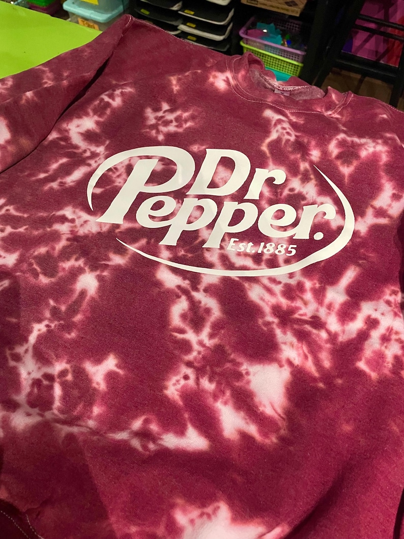 May include: Maroon tie-dye sweatshirt featuring the Dr Pepper logo in white. The logo includes the brand name and "Est. 1885". The sweatshirt has a crew neck and long sleeves. The fabric has a unique, mottled pattern.