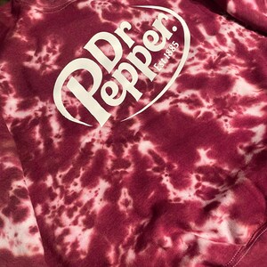 May include: A burgundy tie-dye sweatshirt featuring the Dr. Pepper logo in white. The logo includes the brand name and "Est. 1885". The sweatshirt has a crew neck and long sleeves.