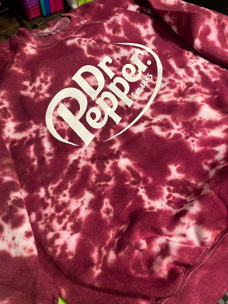 May include: A burgundy tie-dye sweatshirt featuring the Dr. Pepper logo in white. The logo includes the brand name and "Est. 1885". The sweatshirt has a crew neck and long sleeves.