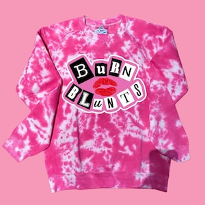 May include: A pink tie-dye sweatshirt with the words "BURN BLUNTS" in a white and black design, featuring a red lip graphic. The sweatshirt is a vibrant pink with white swirls.