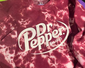 以下が含まれることがあります： 白のDr. Pepperロゴが入った、マルーンのタイダイスウェットシャツ。「Dr Pepper」の文字を様式化したフォントで、下に「Est. 1885」と表示。スウェットシャツはクルーネックと長袖です。