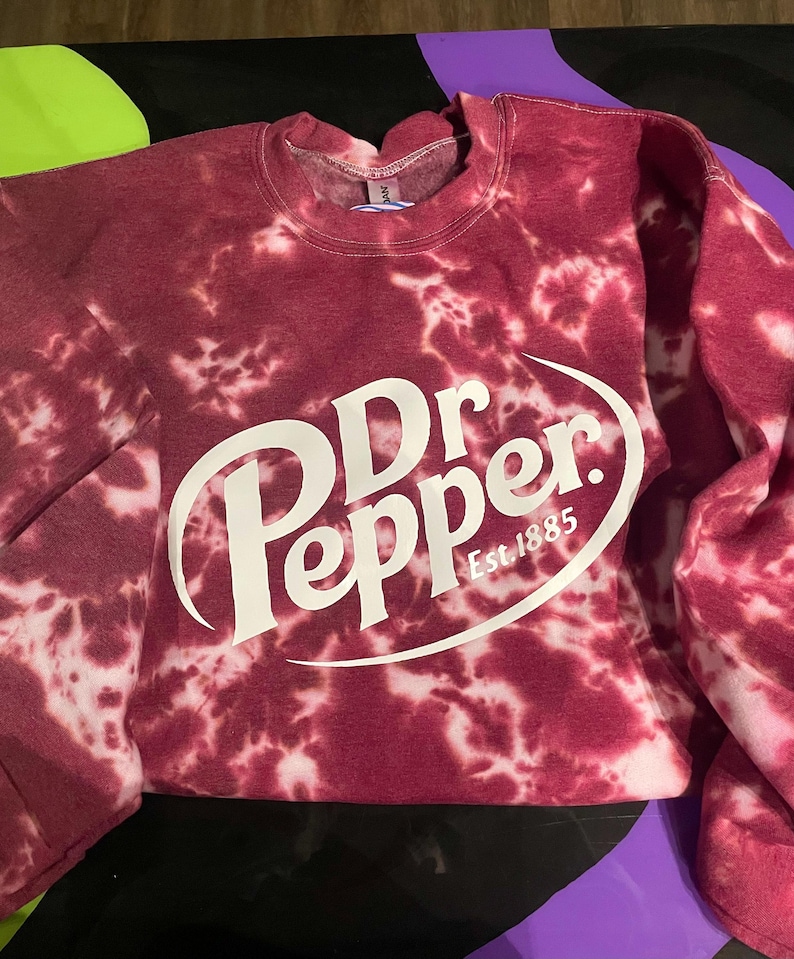 May include: A maroon tie-dye sweatshirt with the Dr. Pepper logo in white. The logo includes the words "Dr Pepper" in a stylized font, with "Est. 1885" below. The sweatshirt has a crew neck and long sleeves.
