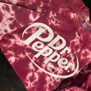 May include: A maroon tie-dye sweatshirt with the Dr Pepper logo in white. The logo includes the words "Dr Pepper" and "Est. 1885" in a white, curved font. The sweatshirt has a crew neck and long sleeves.