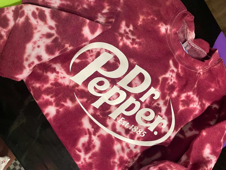 May include: A maroon tie-dye sweatshirt with the Dr Pepper logo in white. The logo includes the words "Dr Pepper" and "Est. 1885" in a white, curved font. The sweatshirt has a crew neck and long sleeves.