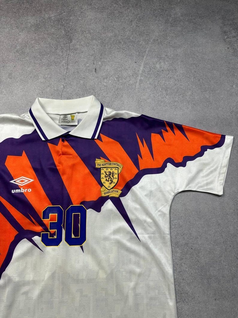 May include: White soccer jersey with orange and purple accents. Features the number 30 in blue, a Scottish Football Association emblem, and the Umbro logo. The jersey has a collared neckline and a geometric design.