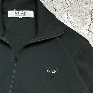 May include: Black zip-up jacket with a high collar. The jacket features a small heart-shaped emblem with eyes on the left chest. The brand name "PLAY COMME des GAR&Ccedil;ONS" is visible on a white tag inside the collar.