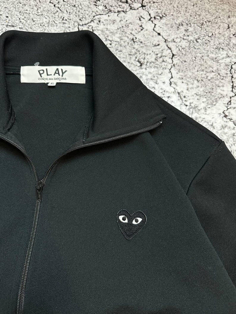 May include: Black zip-up jacket with a high collar. The jacket features a small heart-shaped emblem with eyes on the left chest. The brand name "PLAY COMME des GAR&Ccedil;ONS" is visible on a white tag inside the collar.