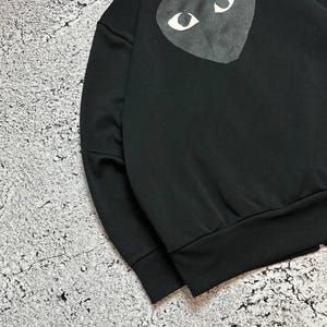May include: Black sweatshirt featuring a large heart-shaped graphic on the back. The graphic has white eyes on a dark gray heart. The sweatshirt has a ribbed hem and cuffs. The collar is a classic style.