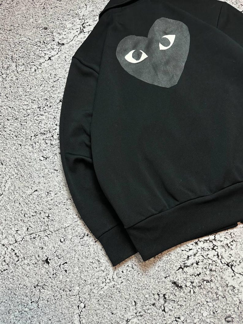May include: Black sweatshirt featuring a large heart-shaped graphic on the back. The graphic has white eyes on a dark gray heart. The sweatshirt has a ribbed hem and cuffs. The collar is a classic style.