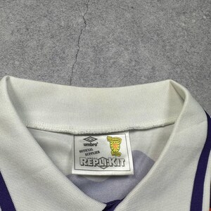 May include: A close-up of a purple and orange soccer jersey with a white collar and accents. The jersey has a tag that reads "Umbro Official Supplier REPLIKIT". The jersey has a white collar and purple and orange stripes.