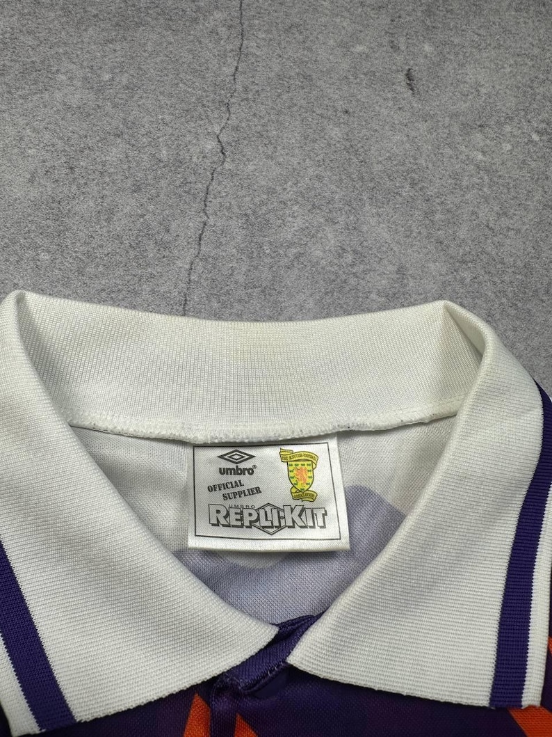 May include: A close-up of a purple and orange soccer jersey with a white collar and accents. The jersey has a tag that reads "Umbro Official Supplier REPLIKIT". The jersey has a white collar and purple and orange stripes.