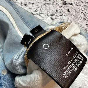 Op de afbeelding: Close-up van een lichtblauwe spijkerbroek met een zwart label. Het label heeft witte tekst met "DRY CLEAN ONLY" en "RN 150712". De jeans heeft zilveren metalen knopen en een maatlabel met "36".