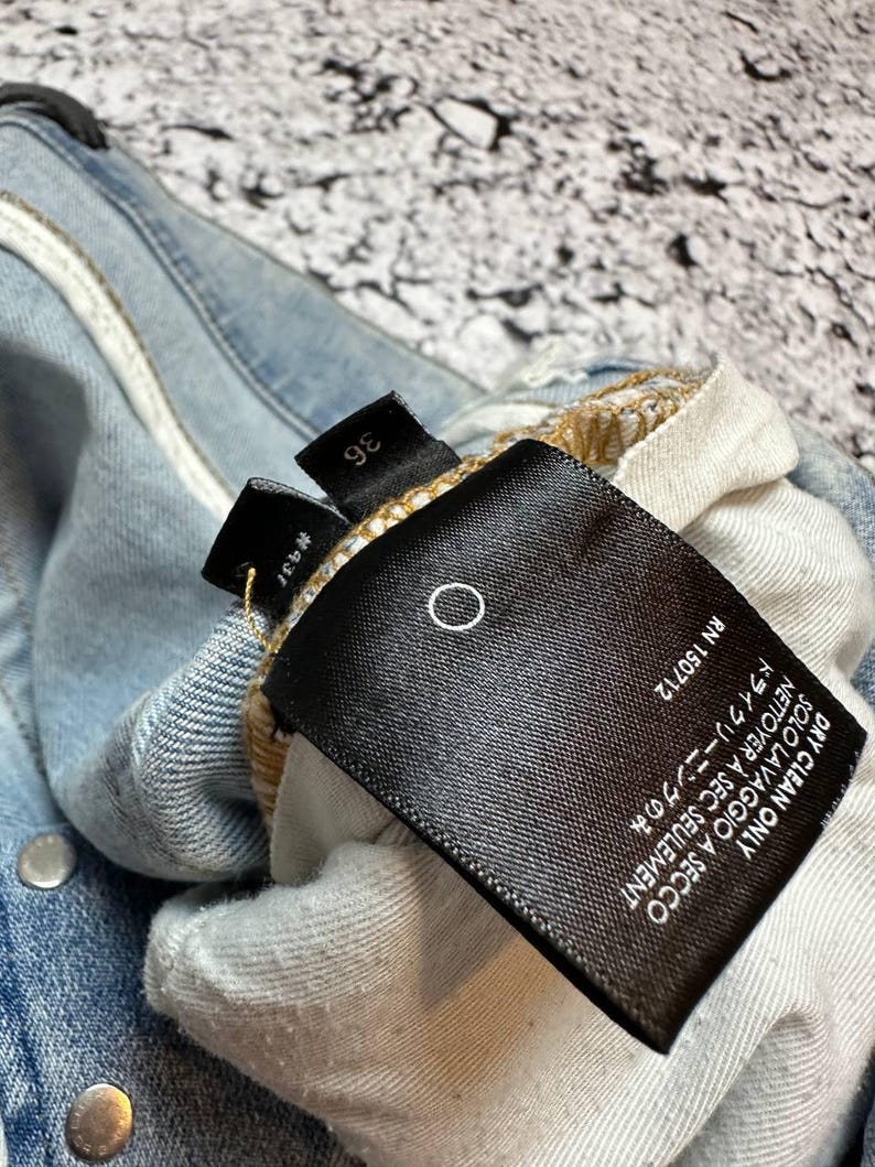 Op de afbeelding: Close-up van een lichtblauwe spijkerbroek met een zwart label. Het label heeft witte tekst met "DRY CLEAN ONLY" en "RN 150712". De jeans heeft zilveren metalen knopen en een maatlabel met "36".