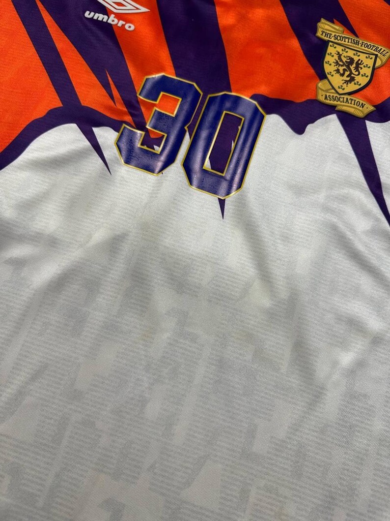 May include: A vintage soccer jersey with a white base, featuring orange and purple accents. The number "30" is prominently displayed in blue with a gold outline. The jersey includes the Umbro logo and a Scottish Football Association emblem.