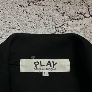 May include: Black garment with a white label that reads "PLAY COMME des GAR&Ccedil;ONS" and "XL". The garment is likely a jacket or shirt, with a visible collar. The background is a textured, light gray surface.