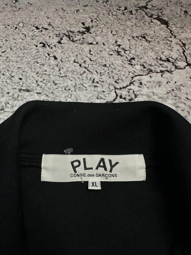 May include: Black garment with a white label that reads "PLAY COMME des GAR&Ccedil;ONS" and "XL". The garment is likely a jacket or shirt, with a visible collar. The background is a textured, light gray surface.