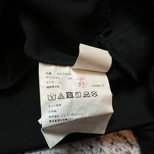 May include: Close-up of a black garment's interior, revealing a white tag with Japanese text and care symbols. The tag includes the product code "AX-T256" and size "XL". The fabric appears to be 100% polyester.