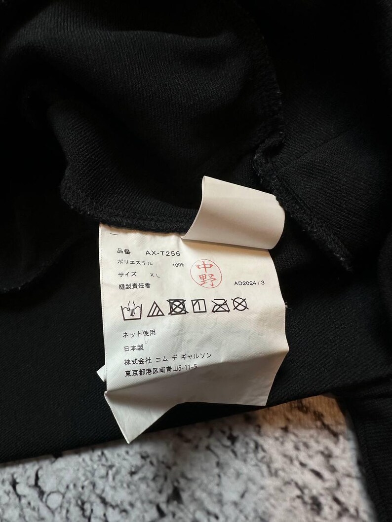 May include: Close-up of a black garment's interior, revealing a white tag with Japanese text and care symbols. The tag includes the product code "AX-T256" and size "XL". The fabric appears to be 100% polyester.