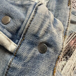 Op de afbeelding: Close-up van een lichtblauwe spijkerjas. De jas heeft zilveren metalen knopen en zichtbare stiksels in een contrasterende geelbruine draad. De stof heeft een versleten, vervaagde uitstraling.