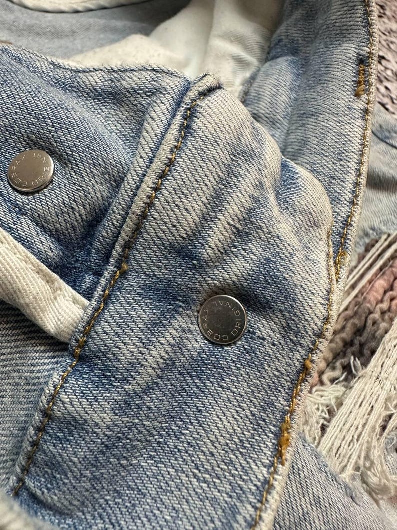 Op de afbeelding: Close-up van een lichtblauwe spijkerjas. De jas heeft zilveren metalen knopen en zichtbare stiksels in een contrasterende geelbruine draad. De stof heeft een versleten, vervaagde uitstraling.