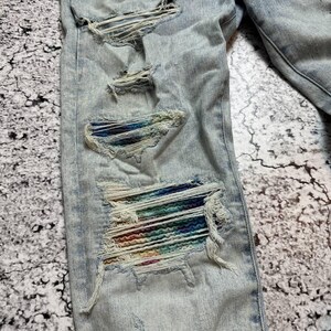 Op de afbeelding: Lichtblauwe denim jeans met distressed details. De jeans hebben meerdere scheuren en gaten, waarvan sommige kleurrijke stof eronder onthullen. De denim heeft een vervaagde uitstraling, met zichtbare stiksels en een klassiek ontwerp met vijf zakken.