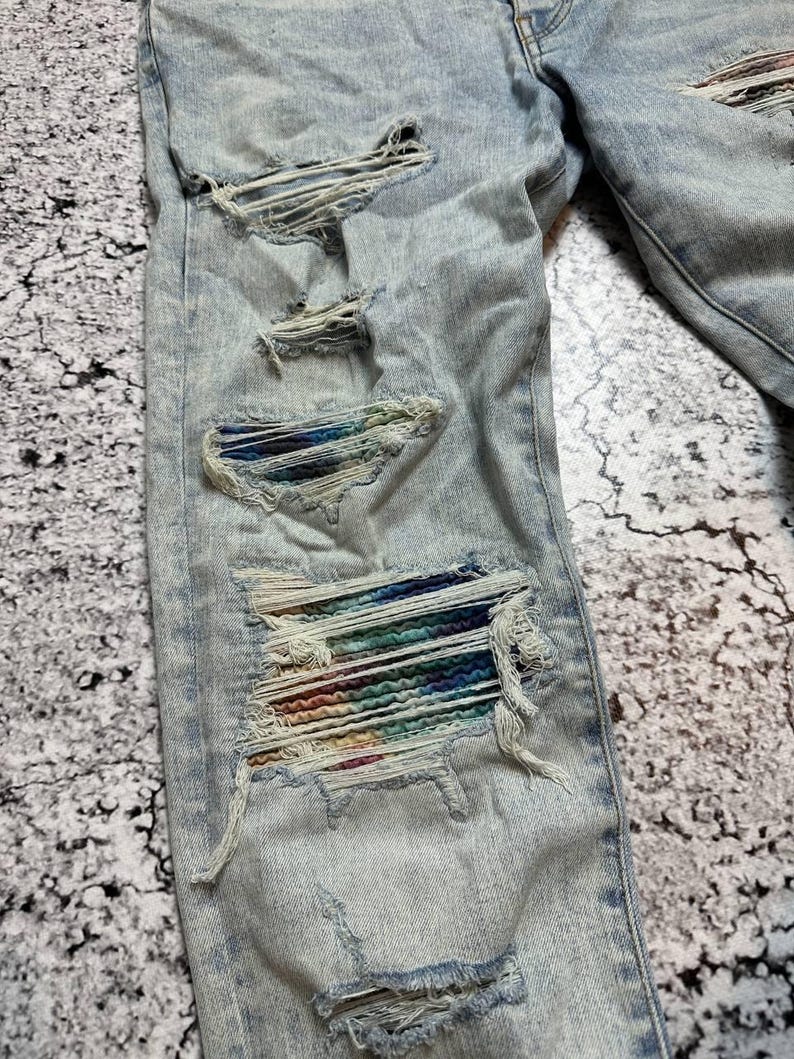 Op de afbeelding: Lichtblauwe denim jeans met distressed details. De jeans hebben meerdere scheuren en gaten, waarvan sommige kleurrijke stof eronder onthullen. De denim heeft een vervaagde uitstraling, met zichtbare stiksels en een klassiek ontwerp met vijf zakken.