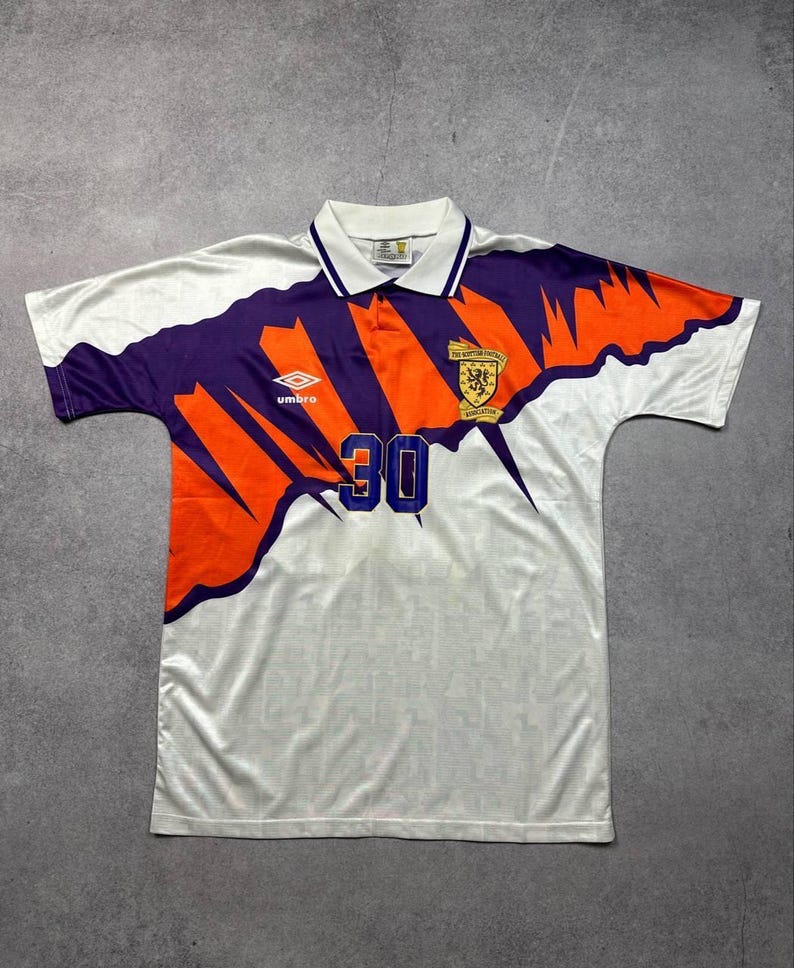 May include: White soccer jersey with purple and orange accents. Features the number 30 in blue, a collar, and the Umbro logo. The jersey has a Scottish Football Association emblem.