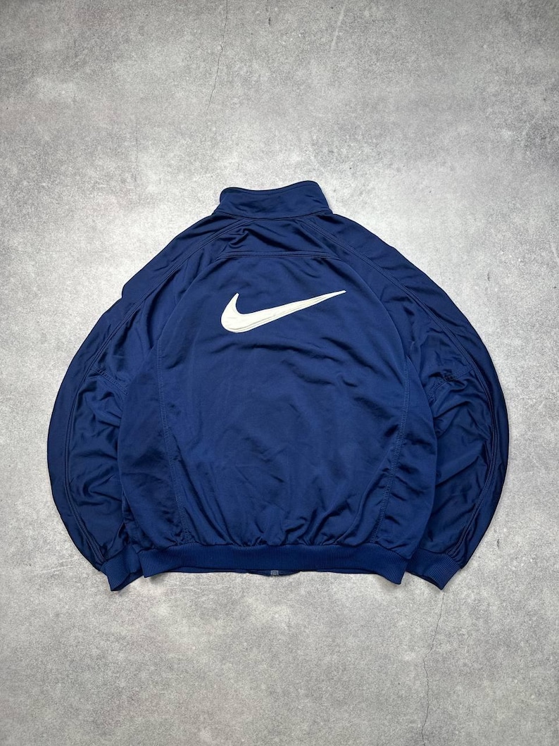 Pode incluir: Casaco de treino azul marinho com o log&oacute;tipo branco da Nike nas costas. O casaco tem fecho de correr completo e gola alta. O tecido &eacute; leve, possivelmente nylon.