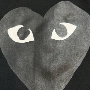 May include: Black garment featuring a large gray heart with white eye shapes. The heart design is centered and takes up most of the frame. The fabric has a subtle texture, and the background is solid black.
