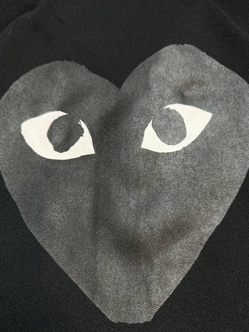 May include: Black garment featuring a large gray heart with white eye shapes. The heart design is centered and takes up most of the frame. The fabric has a subtle texture, and the background is solid black.