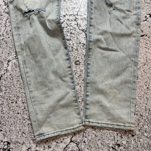 Op de afbeelding: Lichtblauwe denim jeans met distressed details, waaronder scheuren en gerafelde randen. De jeans hebben een kleurrijke, geweven patch bij de bovenbeen. De denim heeft een vervaagde, versleten uitstraling.