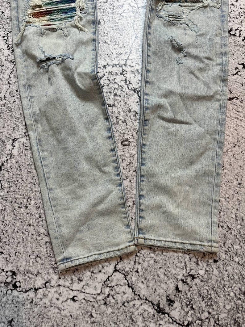 Op de afbeelding: Lichtblauwe denim jeans met distressed details, waaronder scheuren en gerafelde randen. De jeans hebben een kleurrijke, geweven patch bij de bovenbeen. De denim heeft een vervaagde, versleten uitstraling.