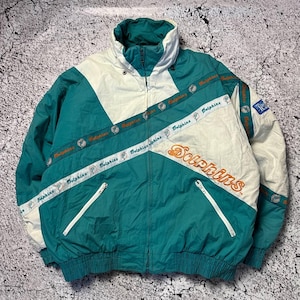 Puede incluir: Chaqueta vintage de color verde azulado, blanco y naranja con la palabra "Dolphins" en escritura naranja. La chaqueta tiene un cuello alto, cierre de cremallera y detalles diagonales blancos. Las mangas tienen una banda blanca con la palabra "Dolphins".