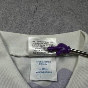 May include: Close-up of a white garment tag with text. The tag reads "85% POLYESTER MINIMUM, L = 108cms, MADE IN ENGLAND". The tag is attached to a white and purple patterned fabric.