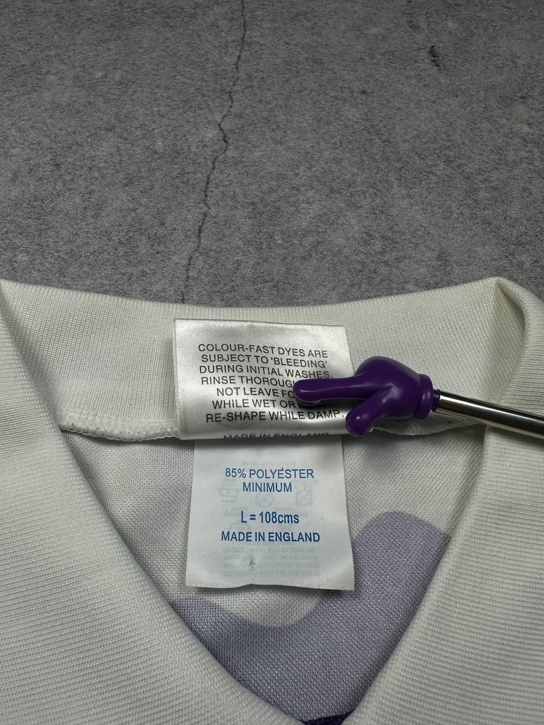 May include: Close-up of a white garment tag with text. The tag reads "85% POLYESTER MINIMUM, L = 108cms, MADE IN ENGLAND". The tag is attached to a white and purple patterned fabric.