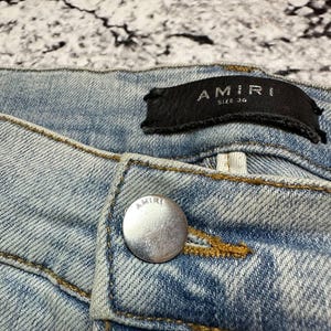 Op de afbeelding: Close-up van lichtblauwe denim jeans met een zilveren knoop en een zwart label. Het label leest "AMIRI SIZE 34". De jeans heeft zichtbare stiksels en een vervaagde wassing.
