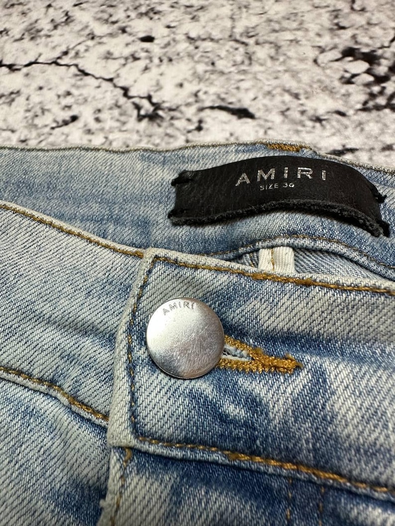 Op de afbeelding: Close-up van lichtblauwe denim jeans met een zilveren knoop en een zwart label. Het label leest "AMIRI SIZE 34". De jeans heeft zichtbare stiksels en een vervaagde wassing.