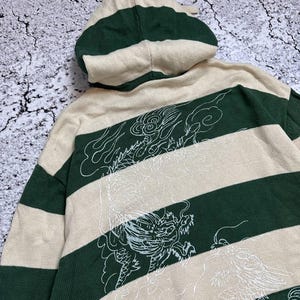 Pu&ograve; includere: Un maglione con cappuccio a righe beige e verdi con un disegno di drago bianco sul retro. Il maglione ha un cappuccio con coulisse e maniche lunghe. Il disegno &egrave; un'illustrazione dettagliata di un drago.