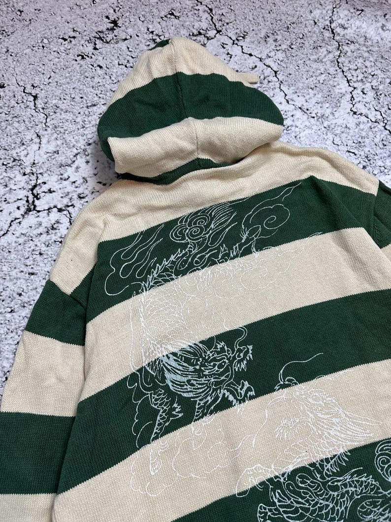 Pu&ograve; includere: Un maglione con cappuccio a righe beige e verdi con un disegno di drago bianco sul retro. Il maglione ha un cappuccio con coulisse e maniche lunghe. Il disegno &egrave; un'illustrazione dettagliata di un drago.